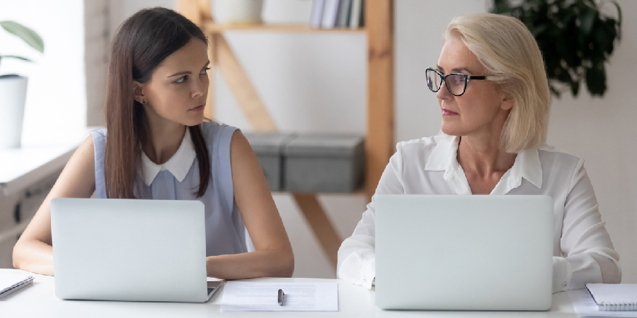 Different ages women colleagues 50s and young female workmates sit at office desk looking with enmity at each other. Negative attitude, conflict at workplace, misunderstanding generational gap concept