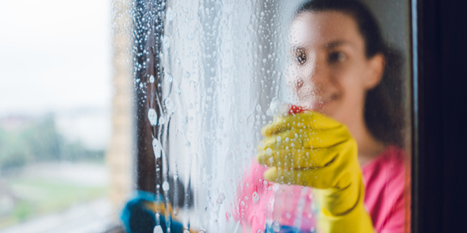 Woman cleaning a mirror, seen in reflection, wearing rubber gloves and spraying blue cleaning fluid