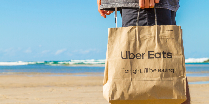 Close up of customer's hand holding Uber Eats paper bag standing on a beach with the water in the background