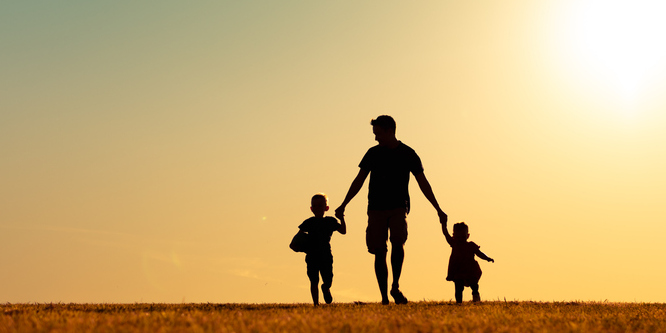 Father holding his children's hands and walking on a beach. The lighting is a sunset and the figures are seen mostly in shadow.