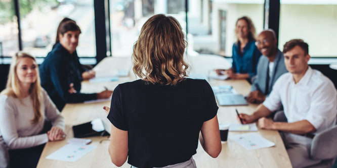 Businesswoman shown from behind in black short-sleeved shirt addressing employees in front of long conference room table