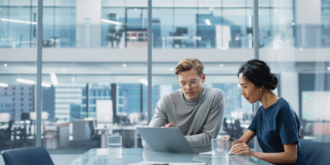 Two younger employees, seated, looking at a laptop in a modern conference room office in front of a large glass window.