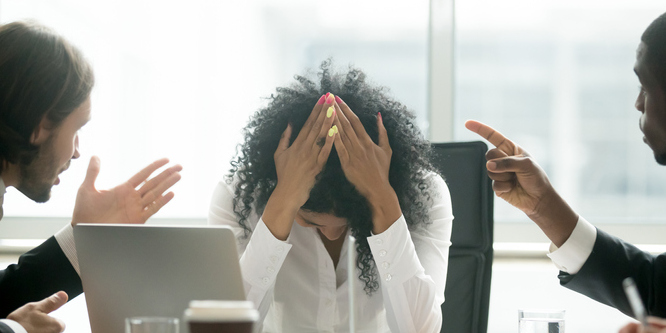 In an office, close up of woman seated in front of a laptop with her head in her hands, being berated by a coworker on either side