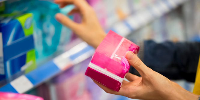 Female hand holding a pink package of female sanitary napkins, other hand browsing a store shelf blurred in the background