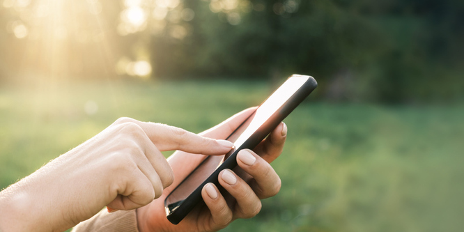 Closeup of hands extending from the side of the photo using a mobile phone, pressing the screen with one index finger, in front of a blurred background of a grassy landscape on a sunny day