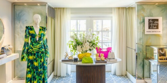 Image of brightly lit room, green patterned dress on manequin to the left, small table with flowers and luxury bags on it in the center in front of large window, jewelry case on the right under sign reading "Saks Fifth Avenue Club"