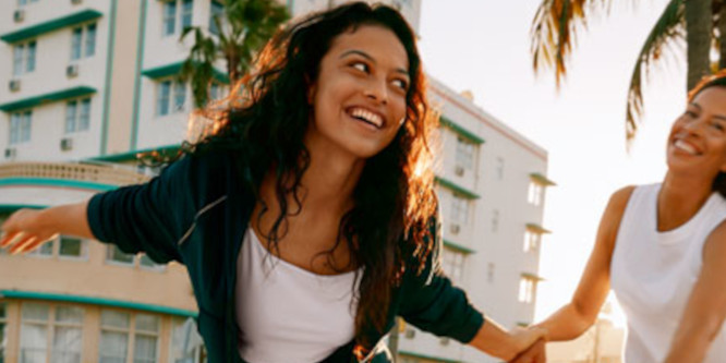 Model and her mother from Victoria's Secret Mother's Day promotion, women holding hands while moving joyfully in front of a white building and a sunny background